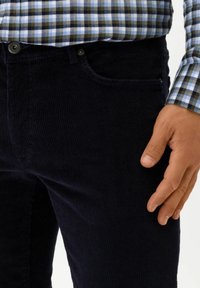 Marina blå cordtöjbyxor med vertikala ribbor, med en framficka och knappstängning, i kombination med en skjorta i blått och grått rutmönster.