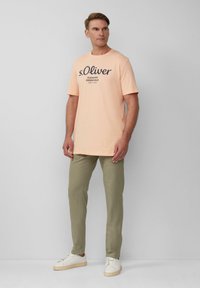 Perzikkleurig katoenen T-shirt met korte mouwen en een marineblauw logo op de voorkant, gecombineerd met olijfgroene broek en witte sneakers.