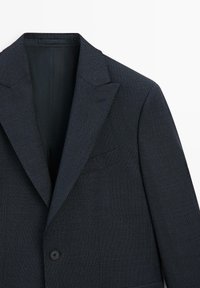 Blazer sartoriale grigio scuro con tessuto strutturato, rever a lancia, chiusura con un pulsante nero singolo e taschino sul petto.
