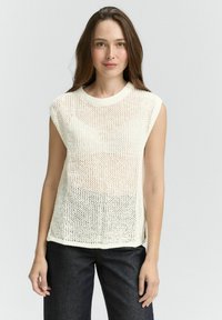 Strickoberteil ohne Ärmel in Creme mit einer lockeren, offenen Webtextur, Rundhalsausschnitt und geradem Saum. Getragen mit dunklen Denim-Hosen.