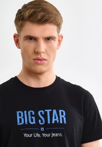 Giovane con capelli castano chiaro corti che indossa una maglietta nera con il testo blu e bianco "BIG STAR Your Life. Your Jeans." su uno sfondo semplice.