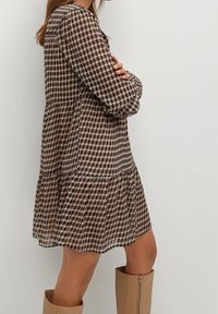 Robe brun à motifs en tissu léger et transparent ; présente un design géométrique, des manches longues et un ourlet évasé.