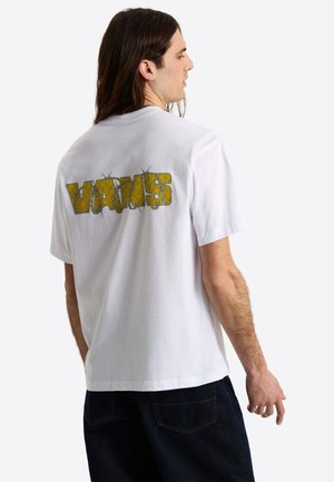 Homme aux longs cheveux foncés portant un T-shirt blanc avec un graphisme jaune style fil barbelé dans le dos et un jean foncé, vu de profil.