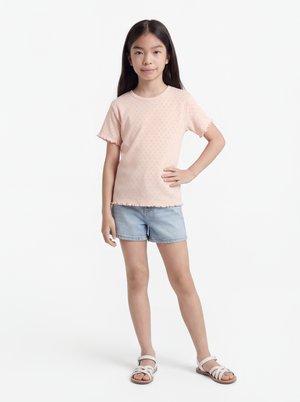 Ung jente står med hånden på hoften, iført lys rosa kortermet topp, denimshorts og hvite sandaler foran en ensfarget hvit bakgrunn.