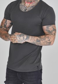 Tmavě šedé tričko s krátkým rukávem a vroubkovaným výstřihem, hladká textura, přiléhavý střih a název značky na lemu.