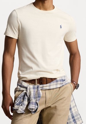 Mand iført en cremefarvet kortærmet t-shirt med lille blåt logo, beige bukser, brun bælte, ur og en blå ternet skjorte bundet om livet.