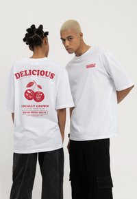 Deux personnes portent des t-shirts blancs surdimensionnés avec le texte rouge "Delicious" sur le thème des fruits et des graphiques, debout devant un fond clair uni.
