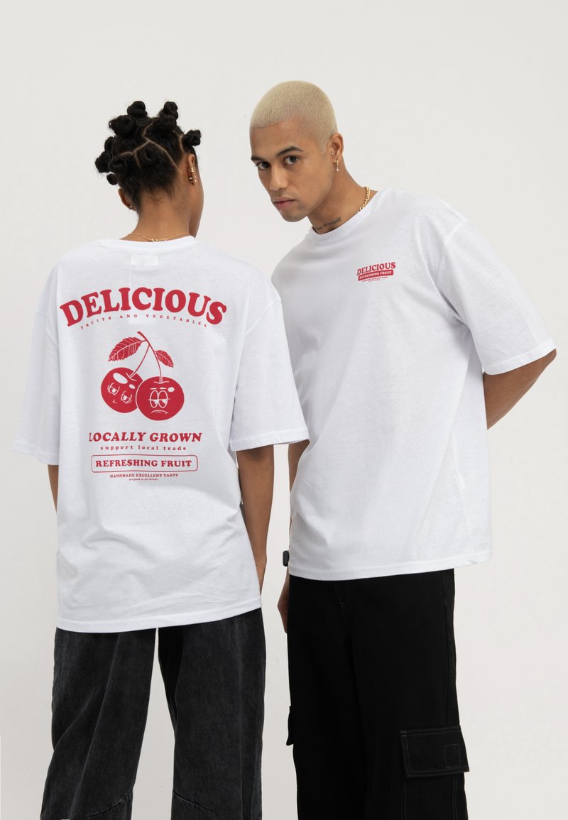 Deux personnes portent des t-shirts blancs surdimensionnés avec le texte rouge "Delicious" sur le thème des fruits et des graphiques, debout devant un fond clair uni.