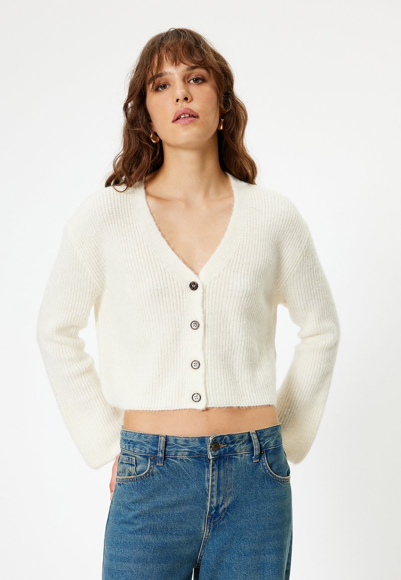Weißer, verkürzter Cardigan mit V-Ausschnitt, gerippter Textur und fünf dunklen Knöpfen an der Vorderseite. Kombiniert mit blauen, hochgeschnittenen Jeans.