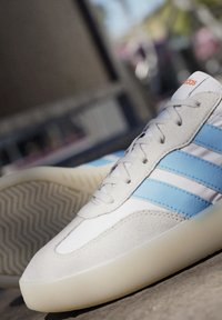 Vita läder och grå mockaskor med blå ränder och orange logotyp, med rund tå och strukturerad gummisula.