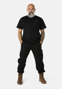 Musta t-paita, löysät mustat cargo-housut Taskuilla ja ruskeat nauhasaappaat. Suunnittelussa on brodeerattu logo paidassa.