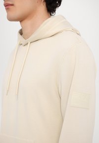 Felpa in cotone beige con cappuccio regolabile, texture liscia e una patch con logo sulla manica sinistra. Design semplice e minimale.