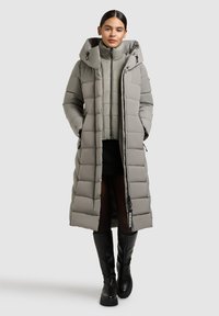 Lange pufferjas in lichtgrijs, met een brede capuchon, een quilted patroon en ritssluiting. Gecombineerd met een cropped binnenjas en hoge zwarte laarzen.