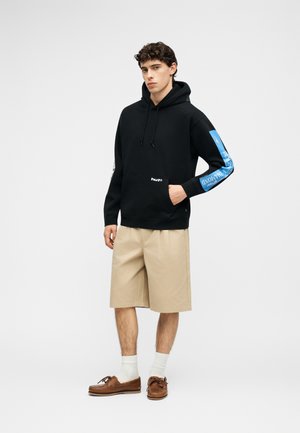 Mladi moški, oblečen v črno kapuco z modrimi besedili na rokavih, bež kratke hlače do kolen, bele nogavice ter rjave čevlje za čoln, stoji pred belim ozadjem.