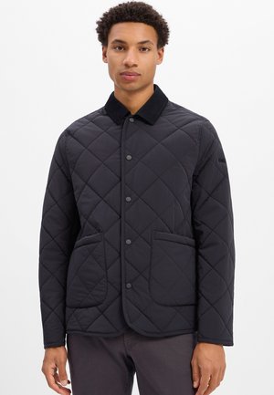 Junger Mann trägt eine schwarze gesteppte Jacke mit Fronttaschen und Druckknöpfen, steht vor einem weißen Hintergrund.