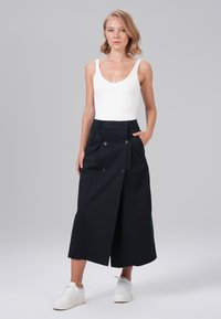 Jupe noire taille haute avec une fente sur le devant, dotée de deux grandes poches et de détails en bouton. Assortie à un body blanc et des baskets blanches.