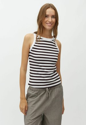 Jeune femme aux longs cheveux bruns portant un top sans manches rayé noir et blanc et un pantalon gris ample, souriante devant un fond blanc uni.