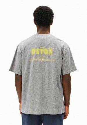 Grijs katoenen t-shirt met korte mouwen. Heeft "DETOX" in geel op de achterkant, met extra tekst daaronder in wit. Losse pasvorm.