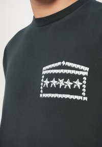 Fekete pulóver, ezüst szegecsekkel díszítve, piramisokkal és csillagokkal a mellkason, kerek nyakkal és texturált anyaggal.