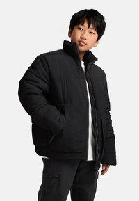 Μαύρο μπουφάν puffer με ψηλό γιακά, κλείσιμο με φερμουάρ και επενδυμένη υφή. Ο σχεδιασμός περιλαμβάνει πλαϊνές τσέπες και άνετη εφαρμογή.