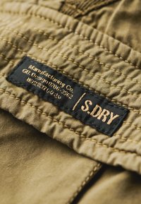 Tkanina khaki o pikowanej fakturze prezentuje czarną metkę z złotym tekstem, na której widnieje napis „Manufacturing Co / S.DRY”, przyszytą do ubrania.