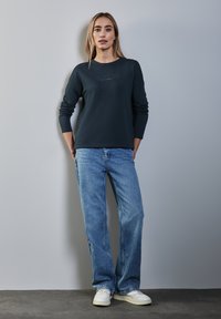 Dunkelblaue Sweatshirt mit Rundhalsauschnitt und gesticktem Logo, kombiniert mit hellblauen Jeans und weißen Sneakern, vor einem neutralen Hintergrund.