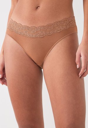 Moteris vilki kūno spalvos medvilninius bikini apatinius su nėriniais, stovinti atsipalaidavusiomis rankomis prie šonų neutraliame fone.