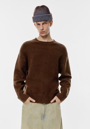 Brązowy, puszysty sweter dzianinowy z okrągłym dekoltem, w połączeniu z jasnoniebieskimi dżinsami. Całość uzupełnia brązowo-niebieska czapka w paski.