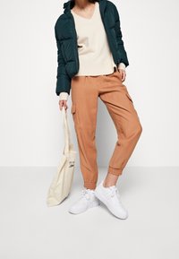 Giacca puffer teal su un maglione crema, abbinata a pantaloni cargo beige e sneakers bianche. Include una borsa a tracolla beige con del testo.