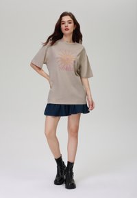 Beige oversized t-skjorte med en solgrafikk og teksten "MYSTICAL SUNRISE." Kombinert med en utslått denimskjørt og svarte ankelstøvler.