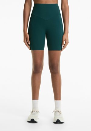 Persoon in donkergroene high-waisted fietsbroek met witte sokken en witte sneakers, staand tegen een witte achtergrond.