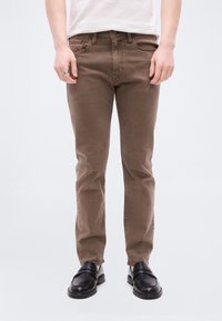 Άτομο που φοράει καφέ στενά τζιν, μαύρα δερμάτινα μοκασίνια και λευκό πουκάμισο, στέκεται μπροστά σε απλό φωτεινό φόντο.