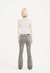Top bianco trasparente a maniche lunghe abbinato a jeans grigi svasati con motivi intricati e dettagli metallici discreti sulle tasche posteriori.