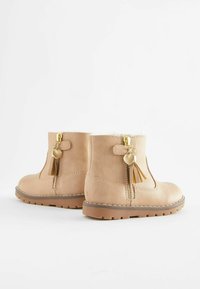 Botas de tobillo de ante beige con cremalleras laterales adornadas con corazones dorados y dijes de borlas, con una suela de goma marrón y costuras blancas.