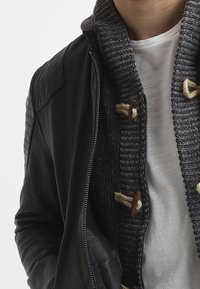 Svart läderjacka med kviltade axlar, lager på lager över en mörk stickad tröja med träknappar och en vit T-shirt under.