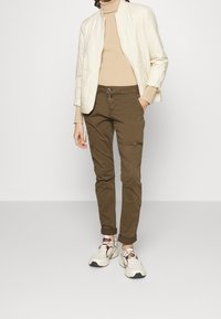 Kvadroderad krämfärgad bomberjacka över en beige turtleneck, matchad med olivgröna byxor och vita sneakers med bruna accenter.