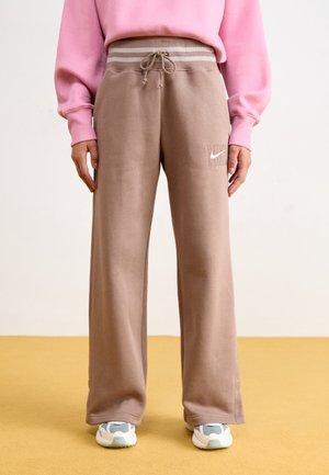 Personne portant un pantalon de survêtement Nike beige à larges jambes avec taille à cordon et des baskets blanches, associé à un sweat-shirt rose.