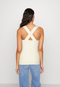 Ribbed tank top in light cream with crisscross back straps and a fitted design. Paired with loose-fitting blue jeans.