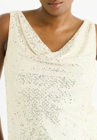 Close-up of a person wearing a sleeveless, cream-colored sequined top with a draped neckline against a white background.