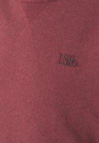Tissu bordeaux avec détail en V cousu et logo LS&Co noir brodé sur la poitrine d’un vêtement.