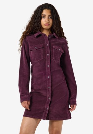 Mulher com cabelo longo e encaracolado vestindo um vestido roxo de veludo cotelê com botões, mangas compridas e bolsos no peito.