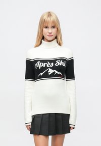 Blond kvinne iført en hvit genser med høy hals med svart fjelldesign og teksten "Après Ski", kombinert med et mørkt plissert skjørt.