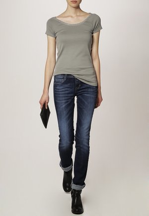 Grijze T-shirt met korte mouwen en geschulpte halslijn, gecombineerd met donkerblauwe, slim-fit jeans. Het model draagt zwarte enkellaarsjes en houdt een zwarte clutch vast.