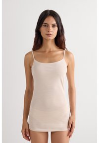 Woman with long dark hair wearing a beige sleeveless dress with thin straps, standing against a plain white background.