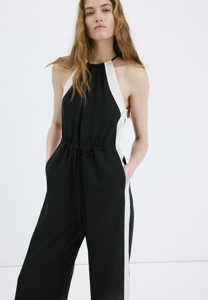 Mujer con cabello largo que lleva un mono negro sin mangas con paneles laterales blancos, de pie con las manos en los bolsillos contra un fondo liso.