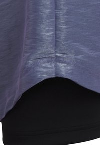 Lila träningsskjorta i ett lättviktigt, texturerat tyg med rundad nederkant. En svart undertröja är delvis synlig nedanför.