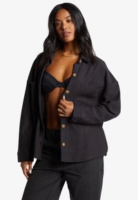 Sort langærmet skjortesjakke lavet af struktureret stof, med store knapper, en brystlomme og en oversized pasform.