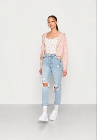 Casaco bomber rosa claro, parte de cima justa branca com estampa floral, jeans claros desgastados, ténis brancos e meias brancas à altura do tornozelo.