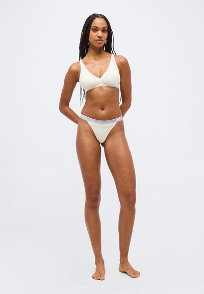 Γυναίκα με μακριά κοτσίδα, ξυπόλυτη, που φοράει μπεζ τοπ μπραζίλιαν τρίγωνο και ταιριαστά σλιπ με λογότυπο στη μέση, σε απλό φόντο.