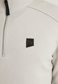 Hellbeige Pullover mit einem halben Reißverschluss, gerippter Textur und einem schwarzen rechteckigen Patch mit orangefarbener Akzentuierung.